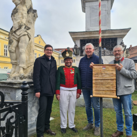 Vier Männer stehen vor einem Denkmal und lächeln in die Kamera. Einer trägt eine Uniform, während die anderen Zettel und Dokumente halten. | © TVB Murau