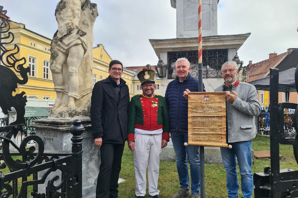 Vier Männer stehen vor einem Denkmal und lächeln in die Kamera. Einer trägt eine Uniform, während die anderen Zettel und Dokumente halten. | © TVB Murau