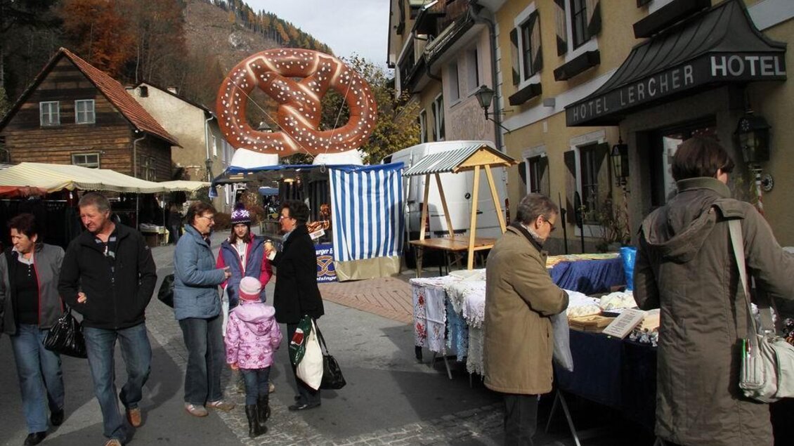 Ein lebhafter Markttag mit Menschen, die entlang der Straße gehen. Stände mit Waren und eine große Brezel als Blickfang im Hintergrund.