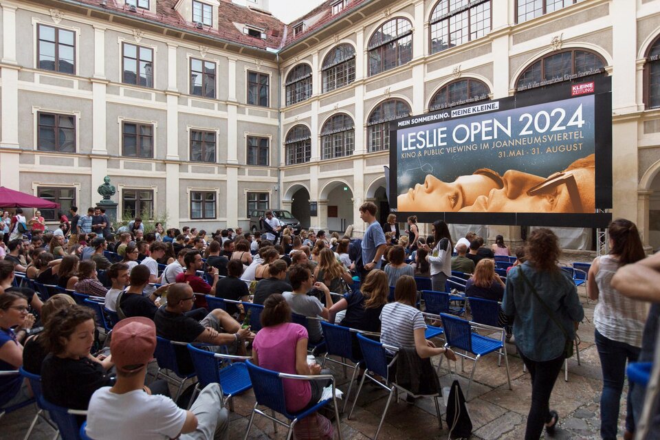 Ein belebter Innenhof mit vielen Menschen, die auf blauem Stuhl sitzen. Im Hintergrund ist ein großes Plakat für die Leslie Open 2024 zu sehen. | © Leslie Open