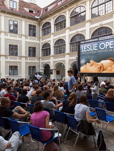 A busy courtyard with many people sitting on blue chairs. In the background, there is a large poster for the Leslie Open 2024. | © Leslie Open