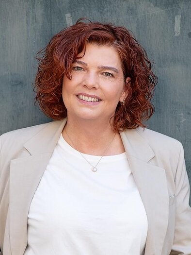 A smiling woman with curly, red hair stands in front of a gray wall. She is wearing a white T-shirt and a beige blazer. | © Tourismus Oststeiermark