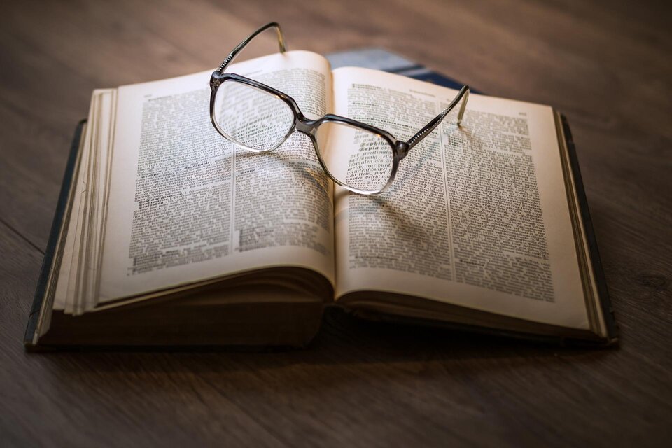 Ein offenes Buch mit Brille darauf liegt auf einem Holztisch. Die Seiten sind mit Text gefüllt.