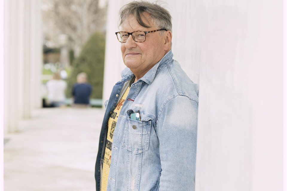 Ein älterer Mann mit Brille steht an einer Wand und lächelt. Er trägt eine Jeansjacke und ein gelbes T-Shirt. | © Lukas Beck