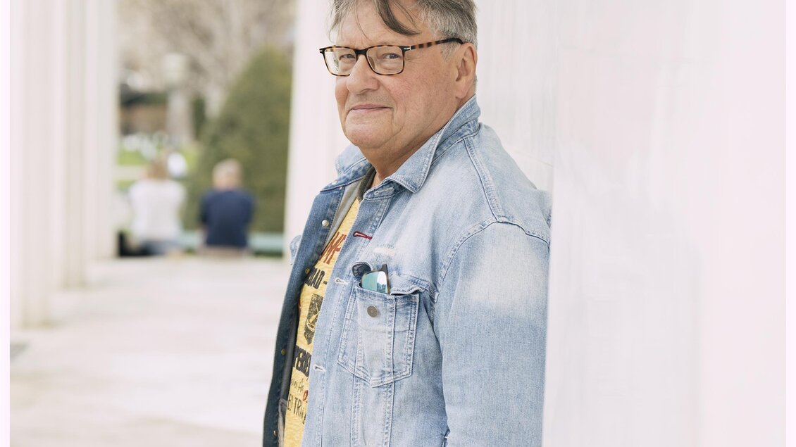 Ein älterer Mann mit Brille steht an einer Wand und lächelt. Er trägt eine Jeansjacke und ein gelbes T-Shirt. | © Lukas Beck