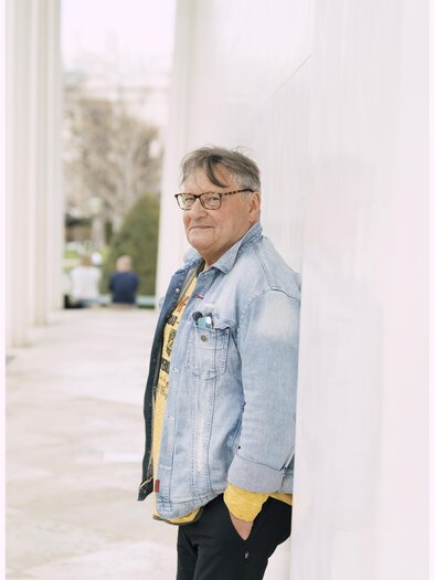 Ein älterer Mann mit Brille steht an einer Wand und lächelt. Er trägt eine Jeansjacke und ein gelbes T-Shirt. | © Lukas Beck