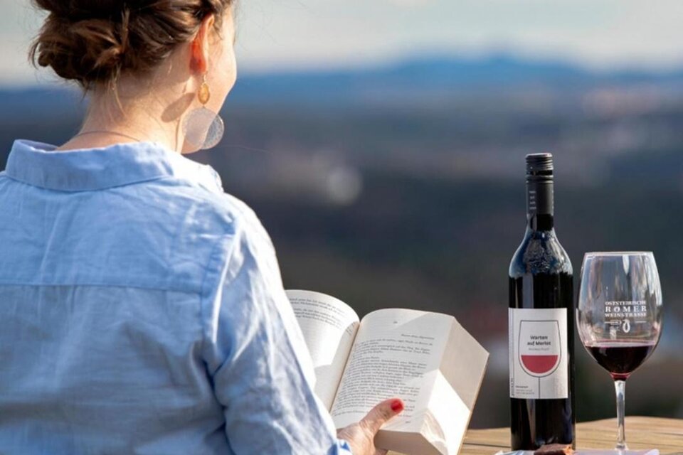 Eine Frau sitzt im Freien und liest ein Buch. Neben ihr stehen eine Flasche Wein und ein Glas Wein, mit schöner Landschaft im Hintergrund. | © Weingut Posch