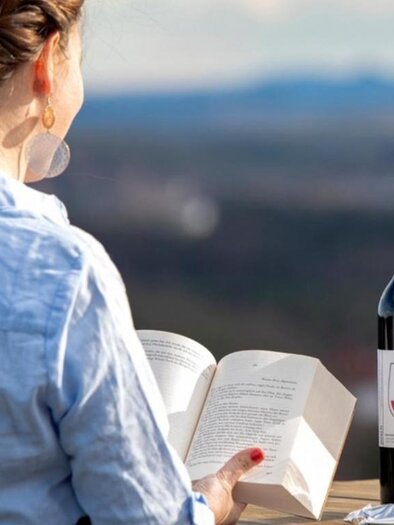 Eine Frau sitzt im Freien und liest ein Buch. Neben ihr stehen eine Flasche Wein und ein Glas Wein, mit schöner Landschaft im Hintergrund. | © Weingut Posch