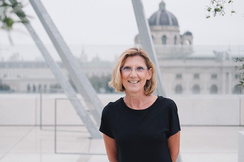 Eine lächelnde Frau steht vor einer modernen Architektur und einem historischen Gebäude im Hintergrund. Die Atmosphäre ist freundlich und einladend. | ©  Günther Wildner
