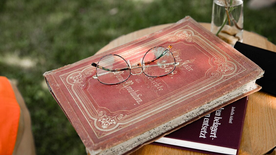 Ein alter, roter Buchdeckel liegt auf einem Tisch, darauf liegen eine Brille und ein weiteres Buch. Im Hintergrund sind grüne Wiesen zu sehen. | © TV Südsteiermark - achromaticphotography