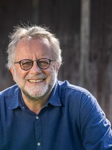 A smiling man with glasses is wearing a blue shirt. In the background, a wooden surface is visible. | ©  Monika Löff