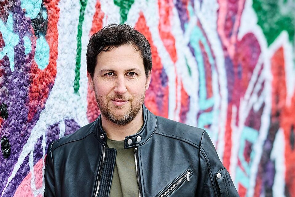 Ein Mann in einer schwarzen Lederjacke steht vor einer bunten Graffiti-Wand. Der Hintergrund zeigt lebendige Farben und kreative Muster. | © René Kovacs