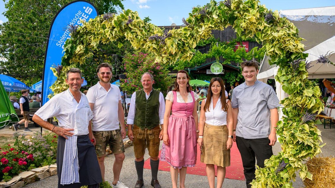 Eine Gruppe von Menschen steht vor einem festlich geschmückten Eingang. Die Personen tragen traditionelle Kleidung und lächeln in die Kamera. | © ©pixelmaker