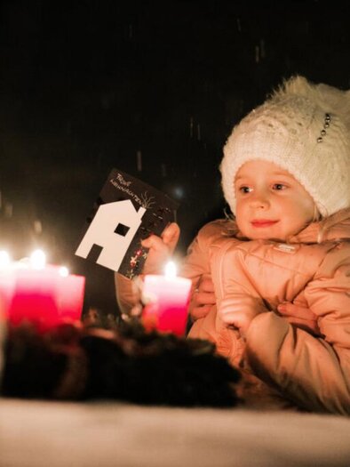 A mother and her small child are sitting at a table in the dark, surrounded by candlelight. Both are wearing warm hats and appear cheerful.