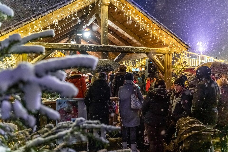 Ein festlicher Weihnachtsmarkt mit vielen Menschen unter einem beleuchteten Holzdach. Schnee bedeckt die Umgebung und schafft eine stimmungsvolle Winteratmosphäre. | © 7000_tv suedsteiermark_stephan friesinger