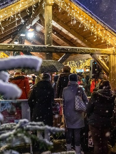 Ein festlicher Weihnachtsmarkt mit vielen Menschen unter einem beleuchteten Holzdach. Schnee bedeckt die Umgebung und schafft eine stimmungsvolle Winteratmosphäre. | © 7000_tv suedsteiermark_stephan friesinger