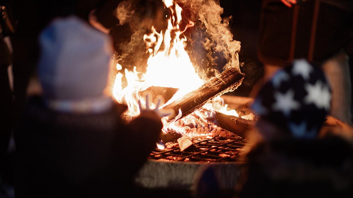Ein warmes Lagerfeuer, das in der Dunkelheit brennt. Zwei Kinder halten ihre Hände in die Nähe der Flammen. | © © TVB Südsteiermark – Achromatic Photography (32)