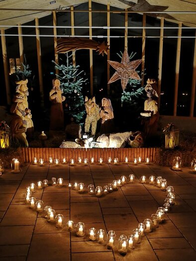 A festive nativity scene with wooden figures stands in a dark room, surrounded by candles. In the foreground, a heart made of candle lights shines. | © Kurkommission Bad Blumau