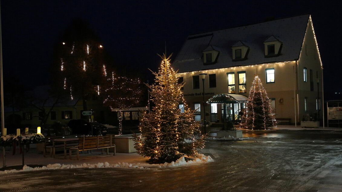 Ein festlich erleuchteter Platz mit geschmückten Weihnachtsbäumen in der Nacht. Im Hintergrund steht ein Gebäude, das ebenfalls weihnachtlich dekoriert ist. | © Kurkommission Bad Blumau