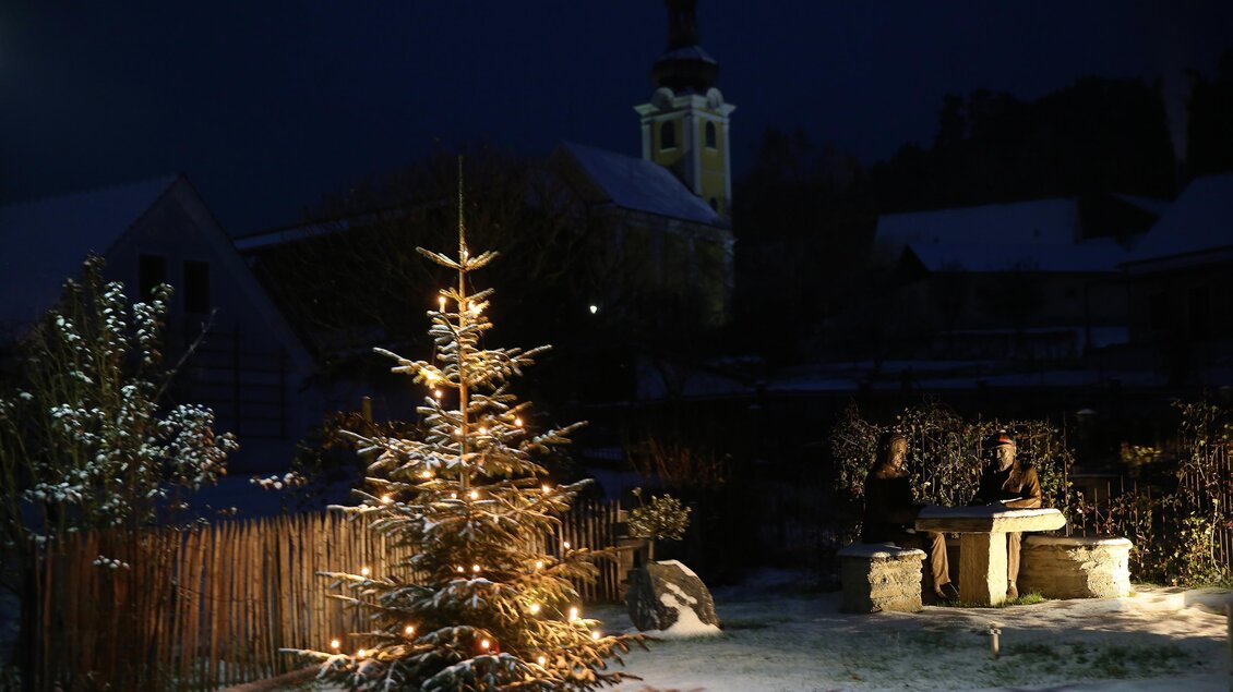 Ein festlich geschmückter Weihnachtsbaum steht im Schnee. Im Hintergrund sind zwei Personen an einem Tisch und eine Kirche zu sehen. | © Kurkommission Bad Blumau