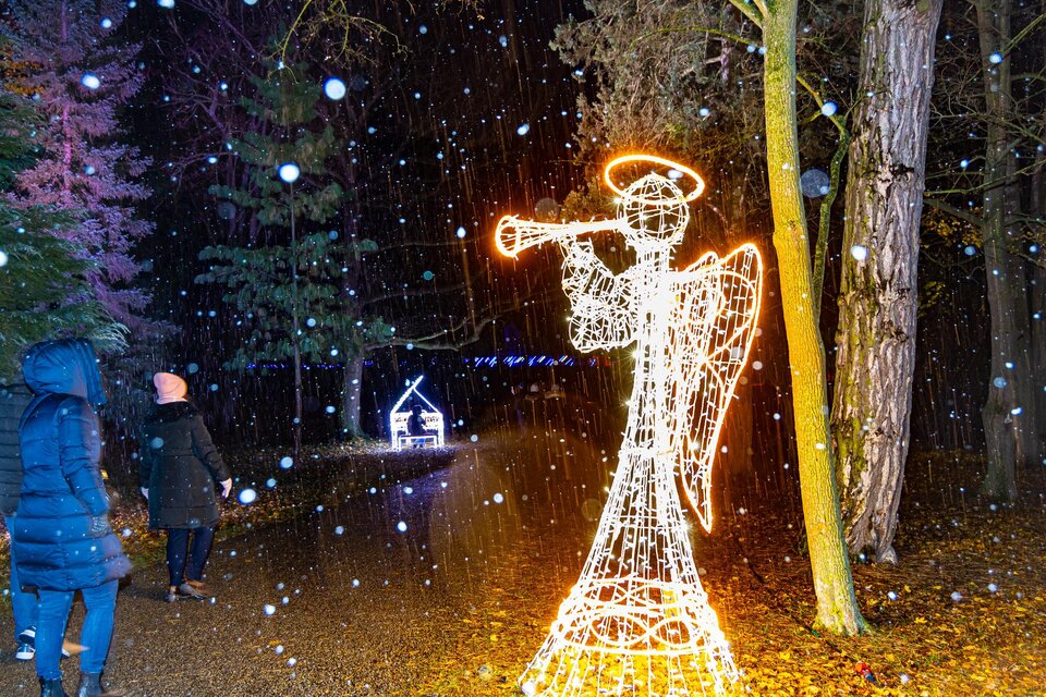 Ein leuchtender Engel mit Trompete steht im Schnee. Zwei Personen spazieren auf einem schmalen Weg, umgeben von Wald. | © © MAGMAG Events GmbH