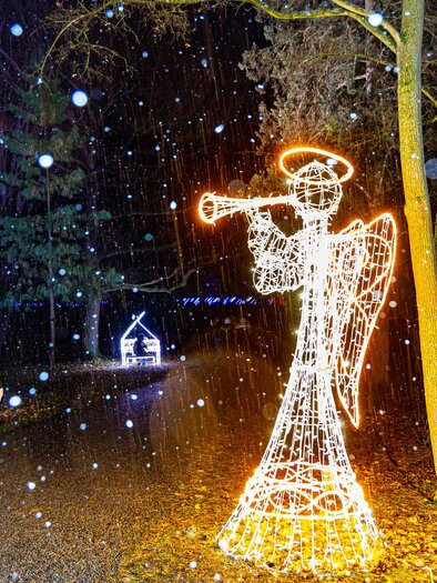 Ein leuchtender Engel mit Trompete steht im Schnee. Zwei Personen spazieren auf einem schmalen Weg, umgeben von Wald. | © © MAGMAG Events GmbH