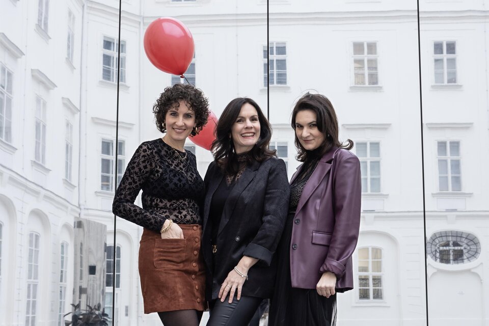 Drei Frauen posieren vor einer gläsernen Wand in einem urbanen Umfeld. Im Hintergrund sind rote Luftballons und elegante Gebäude zu sehen. | © Elisa Maier