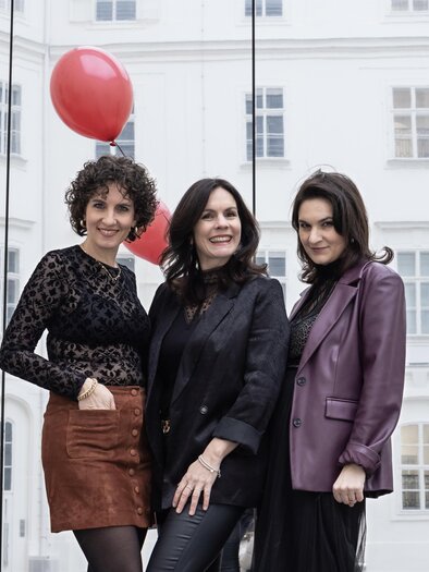 Drei Frauen posieren vor einer gläsernen Wand in einem urbanen Umfeld. Im Hintergrund sind rote Luftballons und elegante Gebäude zu sehen. | © Elisa Maier