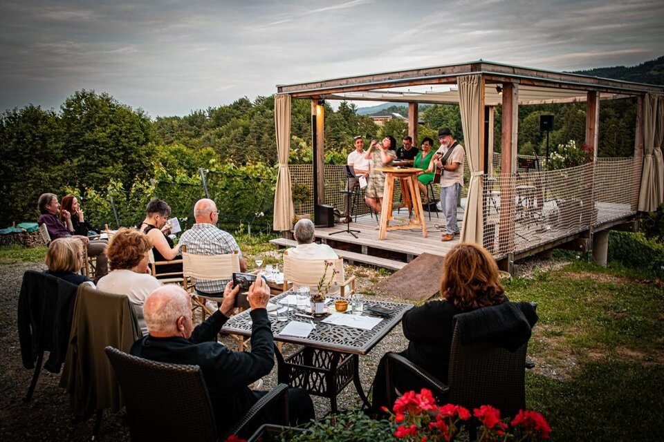 Ein gemütliches Event im Freien mit Gästen, die an Tischen sitzen und einem Live-Auftritt auf einer Terrasse zuschauen. Die Umgebung ist grün und die Atmosphäre entspannt. | © JAsmin Holzmann-Kiefer