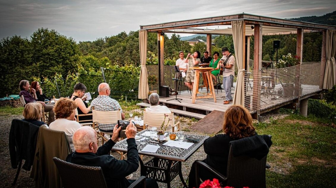 Ein gemütliches Event im Freien mit Gästen, die an Tischen sitzen und einem Live-Auftritt auf einer Terrasse zuschauen. Die Umgebung ist grün und die Atmosphäre entspannt. | © JAsmin Holzmann-Kiefer