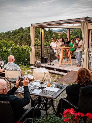 A cozy outdoor event with guests sitting at tables and watching a live performance on a terrace. The surroundings are green and the atmosphere is relaxed. | © JAsmin Holzmann-Kiefer