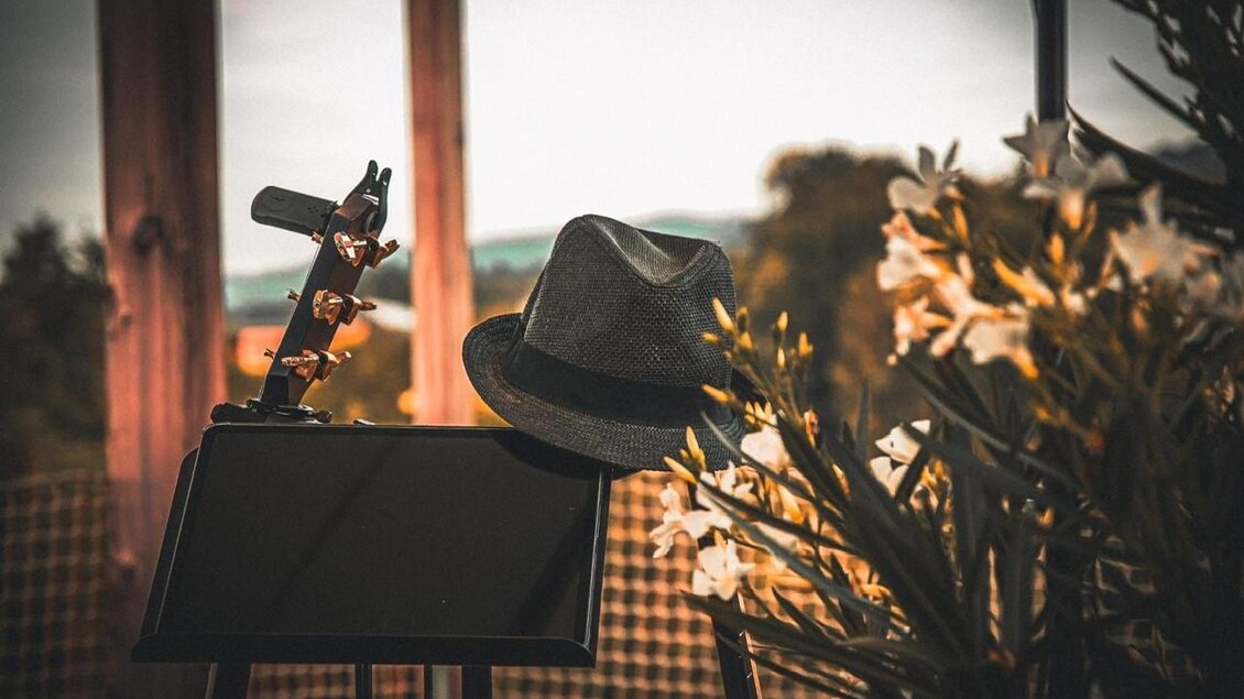 Ein Notenständer mit einer Gitarre und einem Hut steht im Vordergrund. Im Hintergrund sind blühende Pflanzen und eine sanfte Landschaft zu sehen. | © Jasmin Holzmann-Kiefer