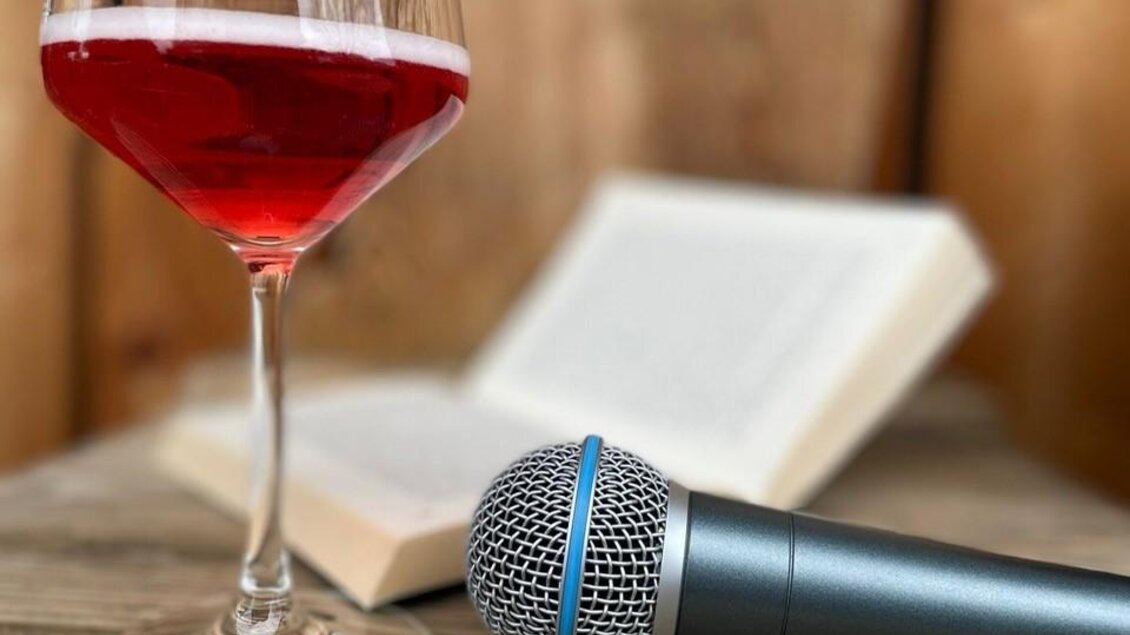 Ein Glas mit rotem Getränk und einem Mikrofon stehen auf einem Holzbrett. Im Hintergrund liegt ein aufgeschlagenes Buch. | © Jasmin Holzmann-Kiefer