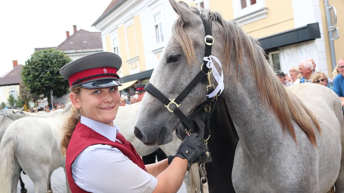 Eine Frau in Uniform steht neben einem schönen grauen Pferd. Im Hintergrund sind Menschen und eine Stadtstraße zu sehen. | © Robert Cescutti