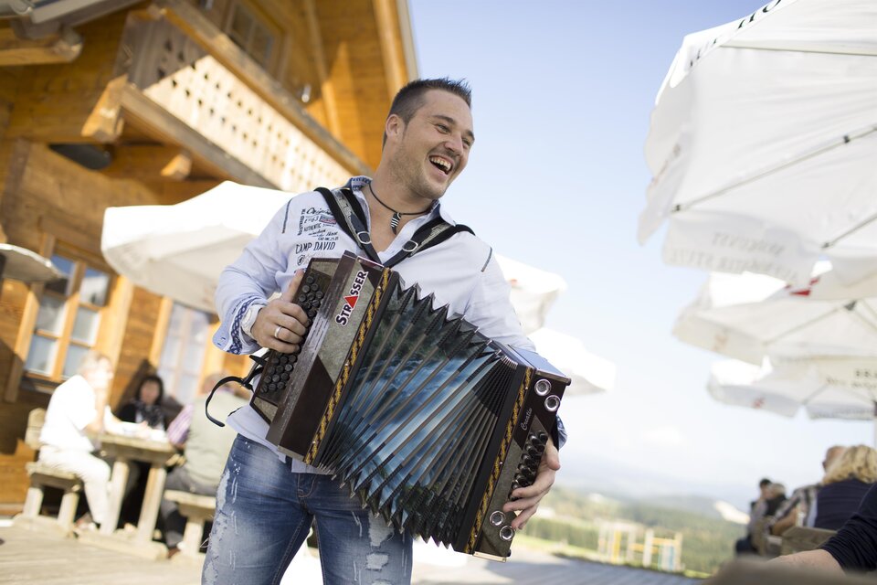 Ein lächelnder Mann spielt Akkordeon vor einem rustikalen Hintergrund. Im Hintergrund sind Sonnenschirme und Menschen zu sehen, die die Atmosphäre genießen. | © BratlAlm