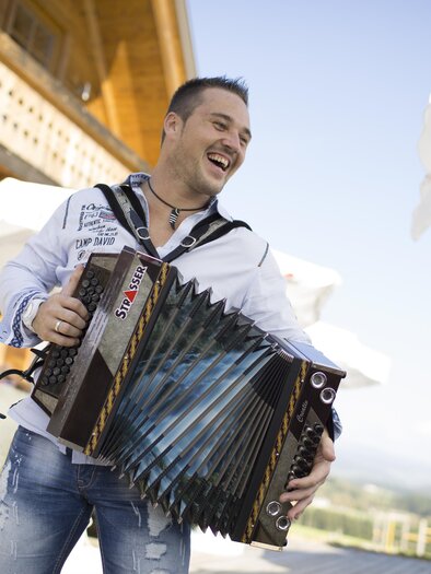 Ein lächelnder Mann spielt Akkordeon vor einem rustikalen Hintergrund. Im Hintergrund sind Sonnenschirme und Menschen zu sehen, die die Atmosphäre genießen. | © BratlAlm