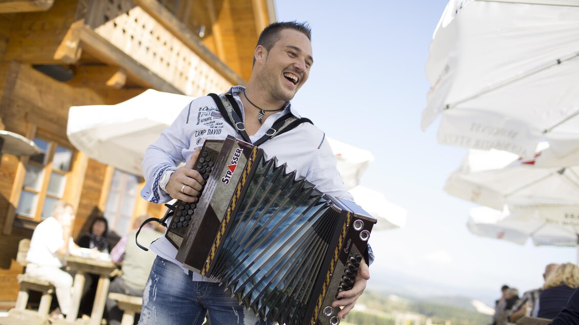 Ein lächelnder Mann spielt Akkordeon vor einem rustikalen Hintergrund. Im Hintergrund sind Sonnenschirme und Menschen zu sehen, die die Atmosphäre genießen. | © BratlAlm