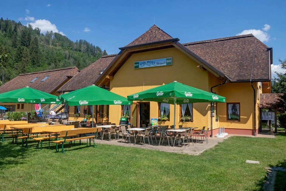 Ein gemütliches Restaurant mit einer gelben Fassade und einem einladenden Außenbereich. Unter grünen Schirmen stehen Tische und Bänke auf einer Wiese, umgeben von Bergen.