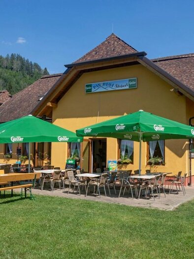 A cozy restaurant with a yellow facade and an inviting outdoor area.  
Under green umbrellas, there are tables and benches on a lawn, surrounded by mountains.
