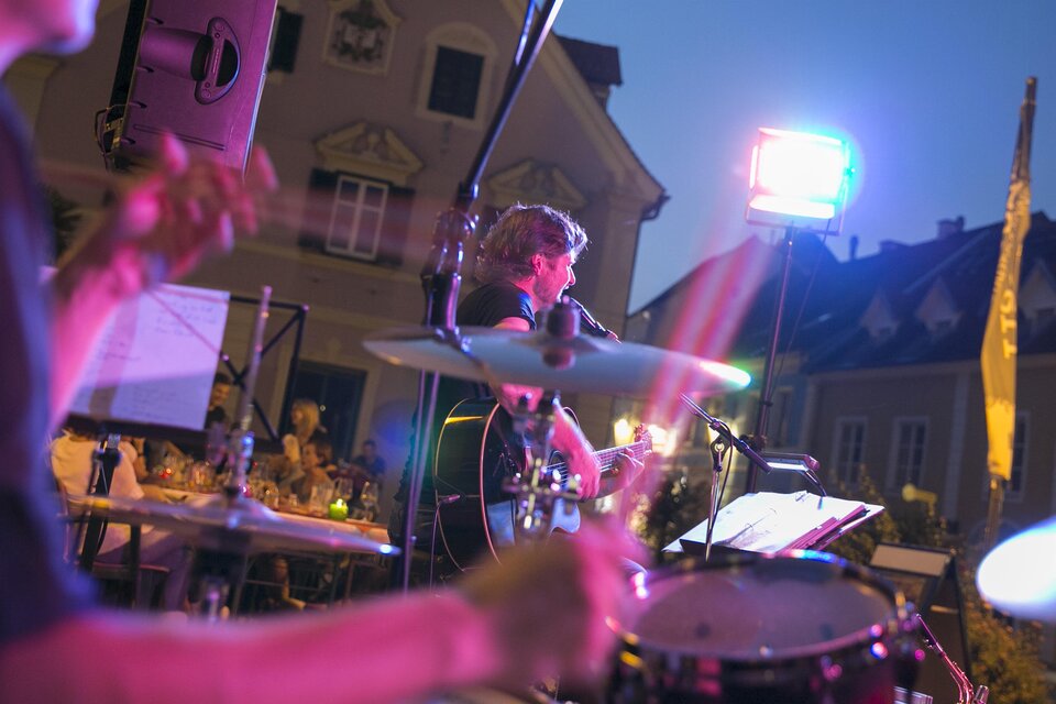 Ein Musiker spielt in der Abenddämmerung auf einer Bühne, während die Bandkollegen im Hintergrund sichtbar sind. Bunte Lichter beleuchten die Szene und schaffen eine festliche Atmosphäre.