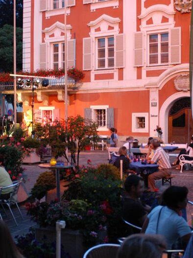 A lively square with guests at a restaurant and colorful flowers. In the background, a historic building and a stage are visible. | © Harald Harmel