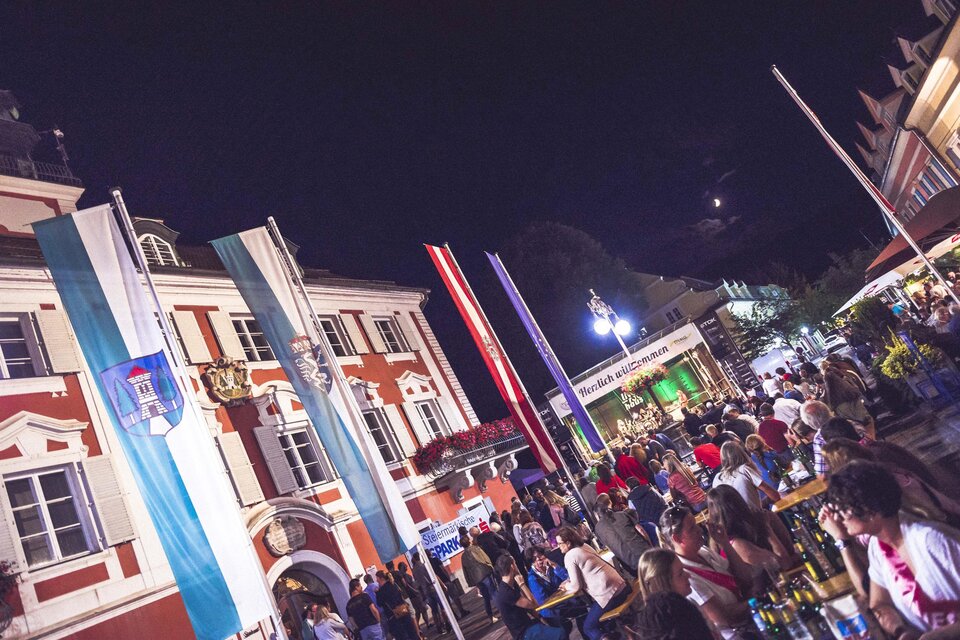 Ein lebhaftes Stadtfest bei Nacht mit vielen Menschen und bunten Fahnen. Im Hintergrund sind festliche Lichter und ein historisches Gebäude zu sehen. | © we shoot it