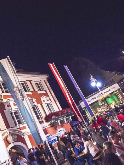 A lively night city festival with many people and colorful flags. In the background, festive lights and a historic building can be seen. | © we shoot it