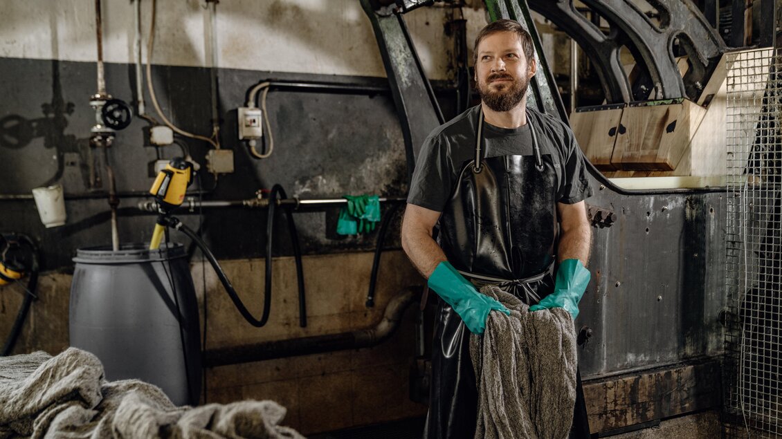 Ein Mann in einem Waschraum steht mit einem Handtuch in der Hand. Er trägt einen schwarzen Schürze und grüne Gummihandschuhe. | © Lodenmanufaktur Steiner1888 | Fotografin Christine Höflehner