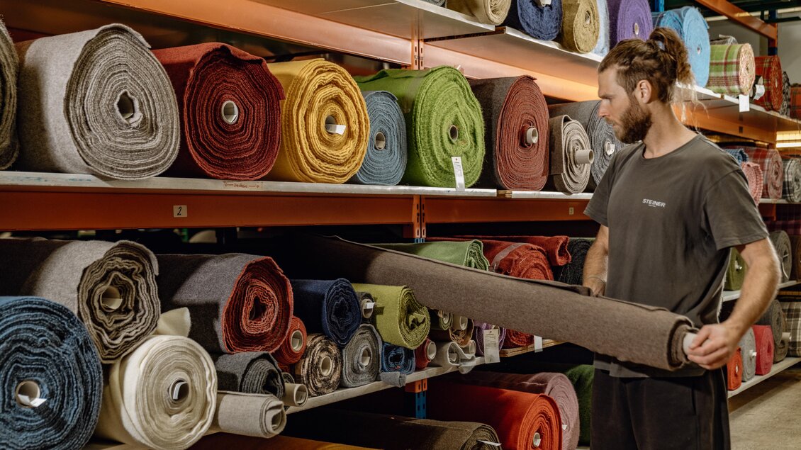 Ein Mann schaut sich Stoffrollen in einem Geschäft an. Die Regale sind mit verschiedenen Farben und Materialien gefüllt. | © Lodenmanufaktur Steiner1888 | Fotografin Christine Höflehner