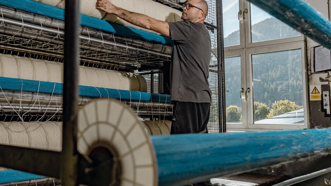 Ein Mann arbeitet in einer Textilfabrik und organisiert Spulen mit Garn. Im Hintergrund sind Fenster, die einen Blick auf die Landschaft bieten. | © Lodenmanufaktur Steiner1888 | Fotografin Christine Höflehner
