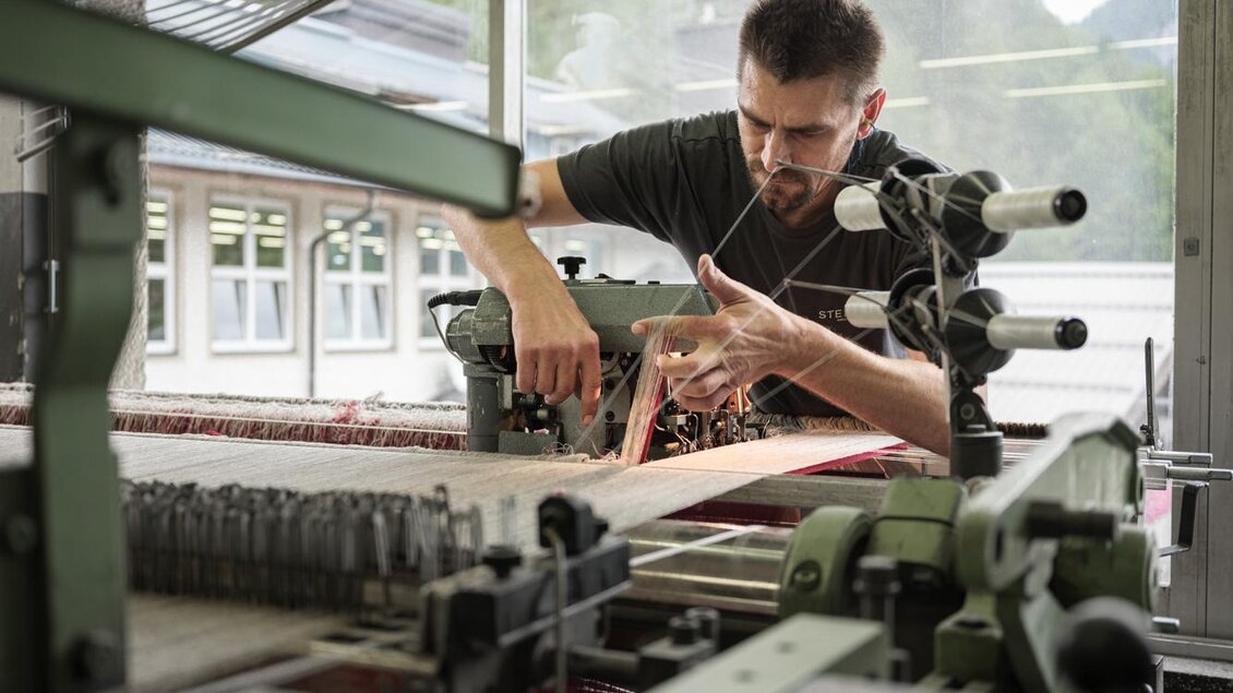 Ein Mann arbeitet an einer Textilmaschine in einer Fabrik. Er konzentriert sich auf die Einstellungen des Geräts, um Stoff herzustellen. | © Lodenmanufkatur Steiner1888 | Fotograf Manuel Schaffernak