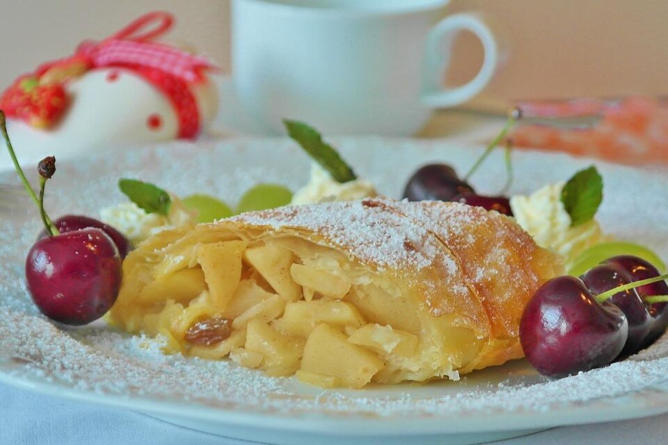 Ein leckeres Stück Apfelstrudel auf einem Teller, garniert mit Kirschen und Minze. Im Hintergrund ist eine Tasse Kaffee sichtbar. | © Pixabay