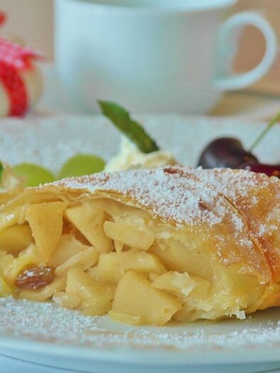 A delicious slice of apple strudel on a plate, garnished with cherries and mint. In the background, a cup of coffee is visible. | © Pixabay
