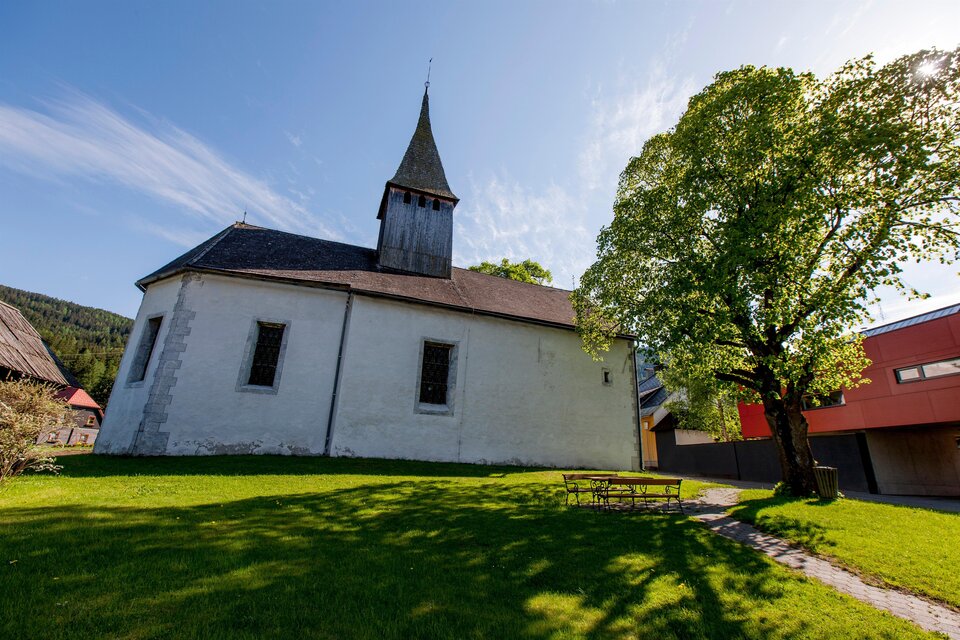 Eine alte Kirche mit einem spitzen Turm und einem großen Baum im Vordergrund. Der Rasen ist grün und die Umgebung wirkt ruhig und einladend.